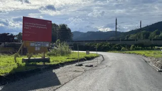 Eine Straße mit einem Schild auf der Seite, das 'Semmering-Basistunnel Unterwerk Langenwang PMZ 5' steht. Die Straße ist leer mit einem Grasbereich auf der Seite. Es gibt einen Zaun mit einem Schild auf der rechten Seite. Bäume und Berge sind in der Ferne.