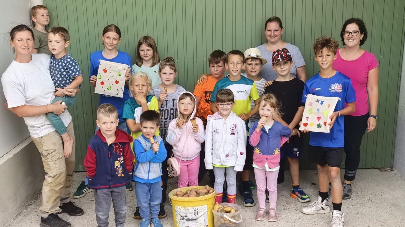 Eine Gruppe von Kindern und einer erwachsenen Frau posiert für ein Foto vor einer grünen Wand und hält äpfelthematische Papiertüten. Zwei Eimer mit Kartoffeln stehen auf dem Boden.