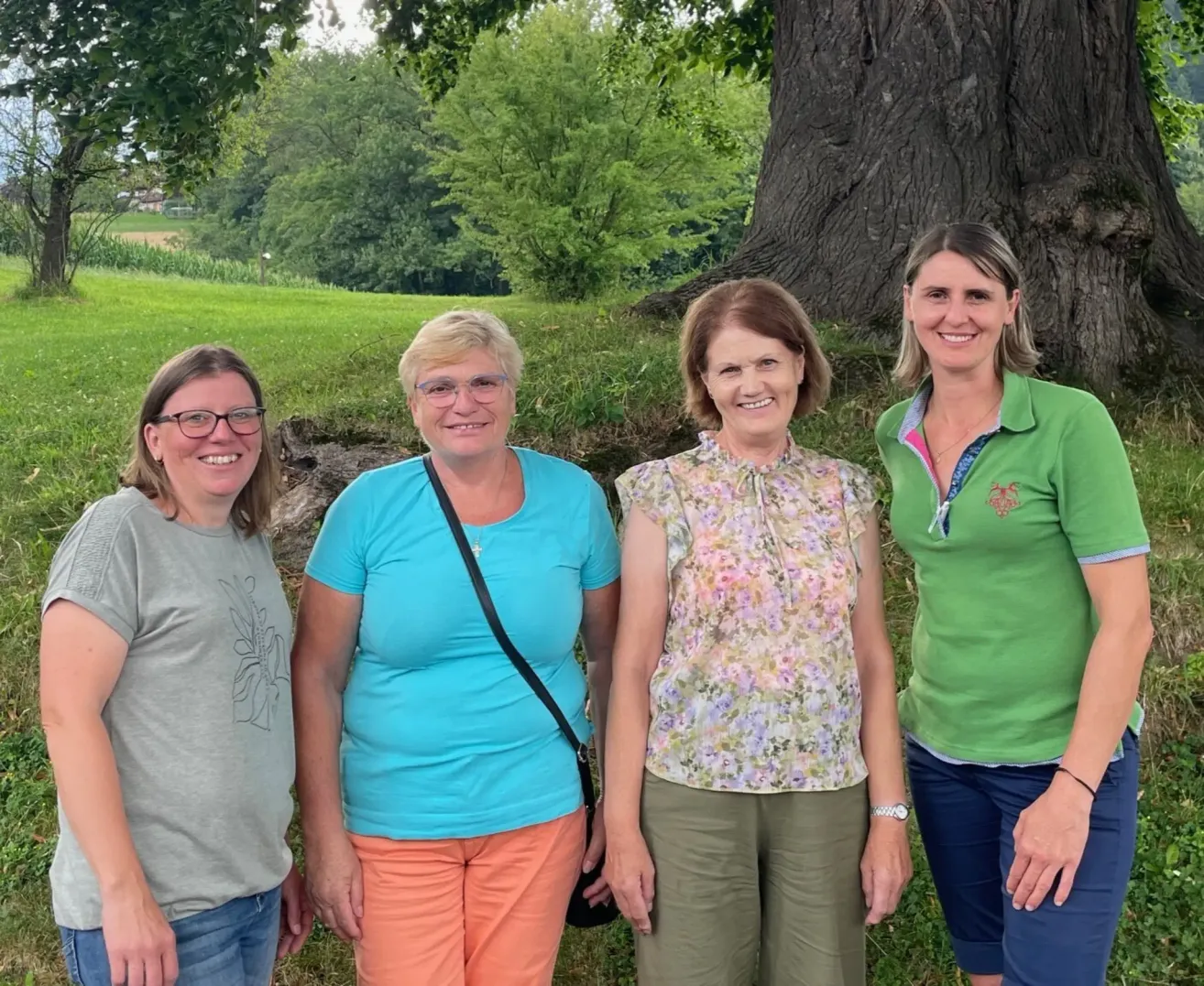 Vier Frauen stehen in einem Park. Sie lächeln alle und posieren für ein Foto vor einem großen Baum.