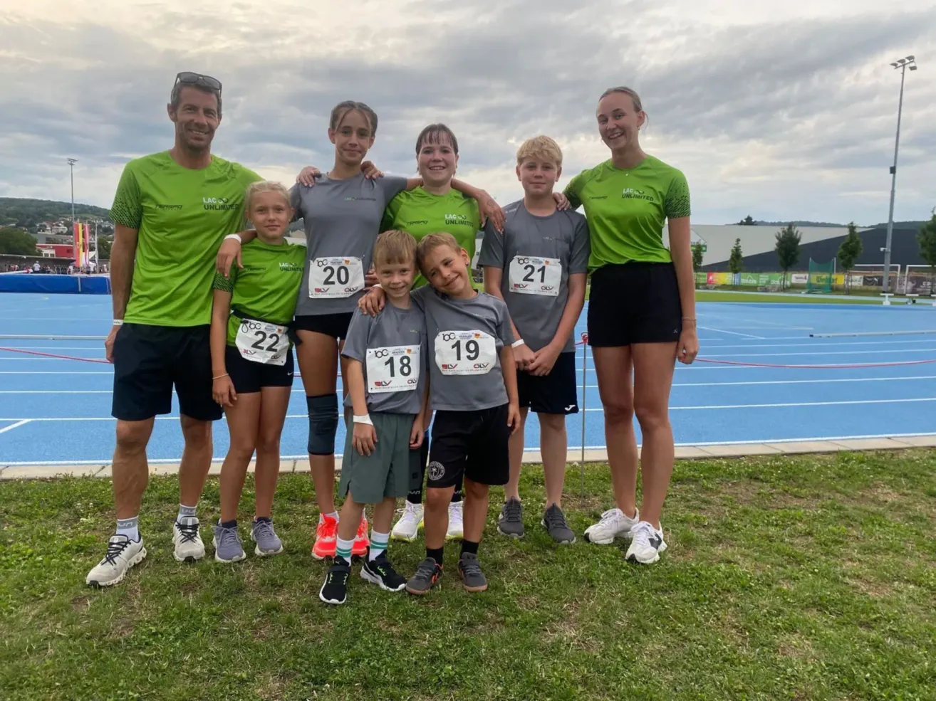 Eine Gruppe von sieben Personen, wahrscheinlich eine Familie, steht zusammen auf einem Rasenfeld, alle tragen passende Shirts mit Startnummern. Im Hintergrund ist eine Laufbahn zu sehen.