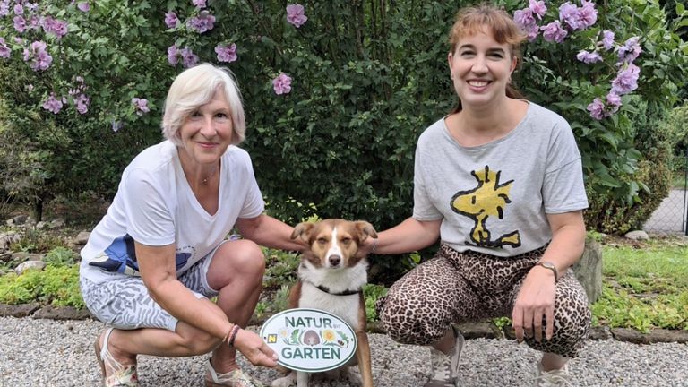 Zwei Frauen und ein Hund posieren für ein Foto in einem Garten. Sie lächeln und halten ein Schild mit der Aufschrift 'Natur Garten'. Die Frau auf der linken Seite trägt ein Armband.
