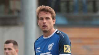 Ein Mann in einem blauen Fußballtrikot mit der Nummer 19 schaut etwas vor sich an.