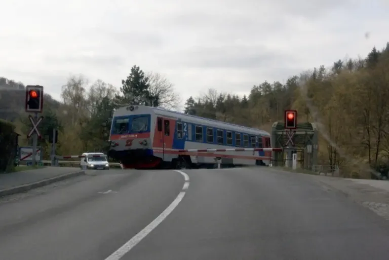 Ein Zug mit der Nummer 2 überquert eine Kreuzung. Eine rote Ampel signalisiert Stopp. Ein weißer Lieferwagen ist am Straßenrand geparkt.