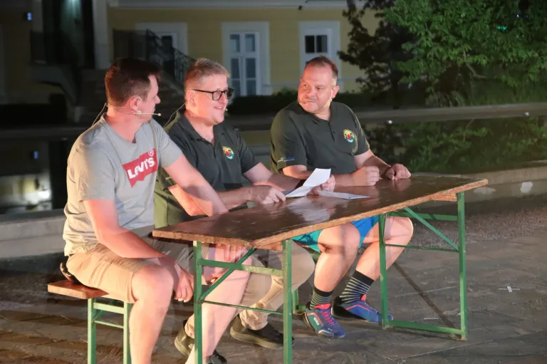 Drei Männer sitzen draußen um einen Tisch herum, einer trägt ein Levi's-Shirt, ein anderer eine Brille und der dritte hält ein Papier. Sie scheinen ein Gespräch zu führen.
