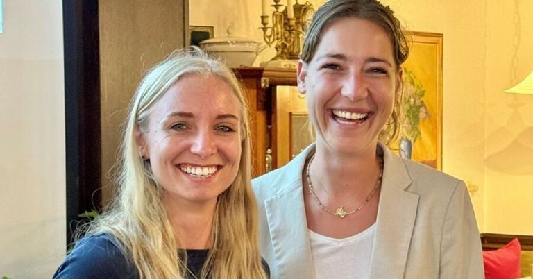 Zwei Frauen, eine mit blonden Haaren und die andere mit braunen, lächeln und posieren für ein Foto in einem Raum mit einem goldenen Kronleuchter.