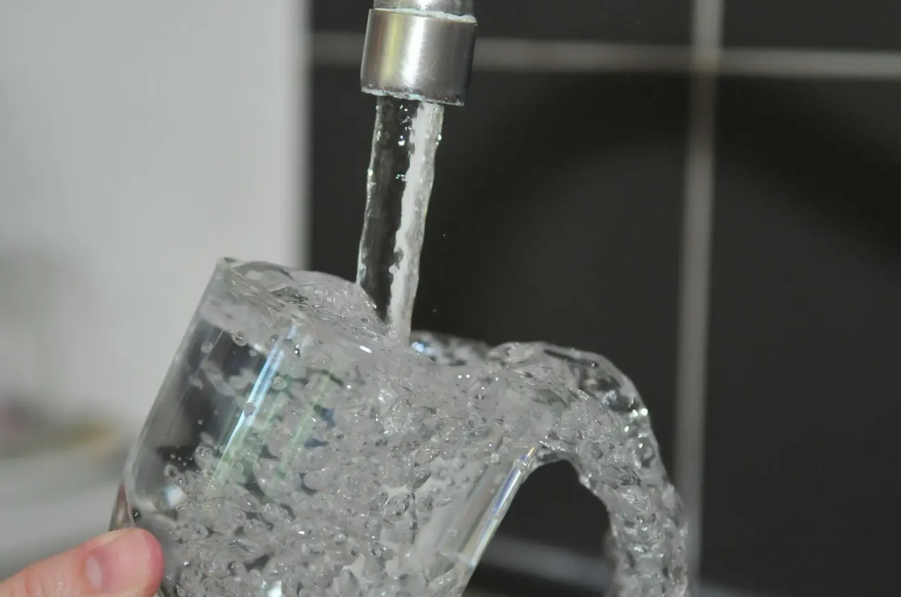Wasser wird aus einem Wasserhahn in ein klares Glas gegossen. Das Wasser fließt gleichmäßig und spritzt leicht.