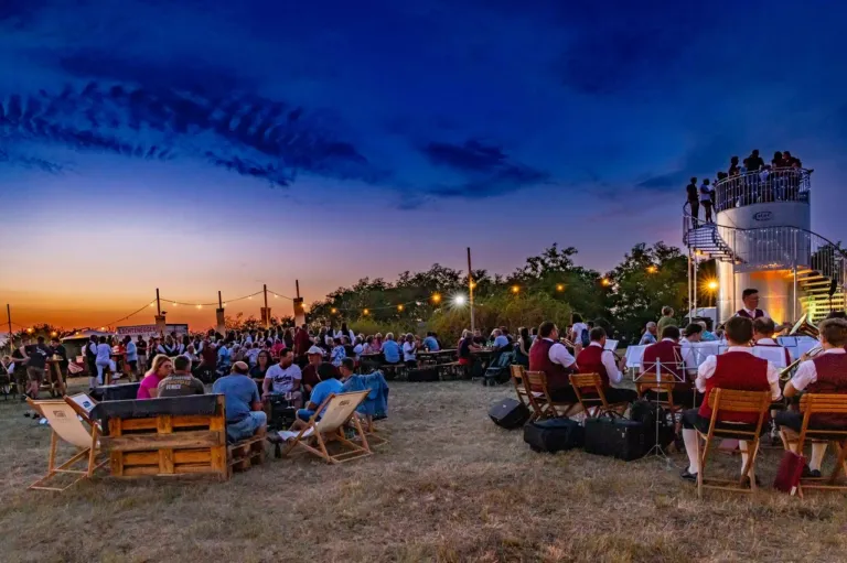 Eine Outdoor-Veranstaltung bei Dämmerung mit vielen Menschen, die auf Stühlen und Tischen sitzen. Eine Band spielt auf einer Bühne, und einige Leute stehen auf dem Balkon. Der Himmel ist blau mit einigen Wolken, und es gibt Bäume und Straßenlaternen in der Umgebung.