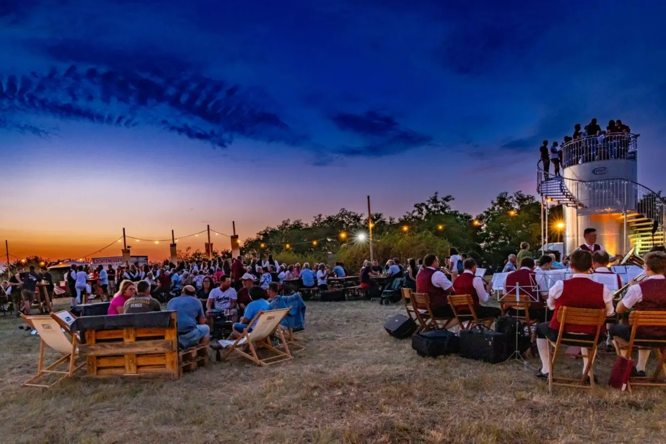 Eine Outdoor-Veranstaltung bei Dämmerung mit vielen Menschen, die auf Stühlen und Tischen sitzen. Eine Band spielt auf einer Bühne, und einige Leute stehen auf dem Balkon. Der Himmel ist blau mit einigen Wolken, und es gibt Bäume und Straßenlaternen in der Umgebung.