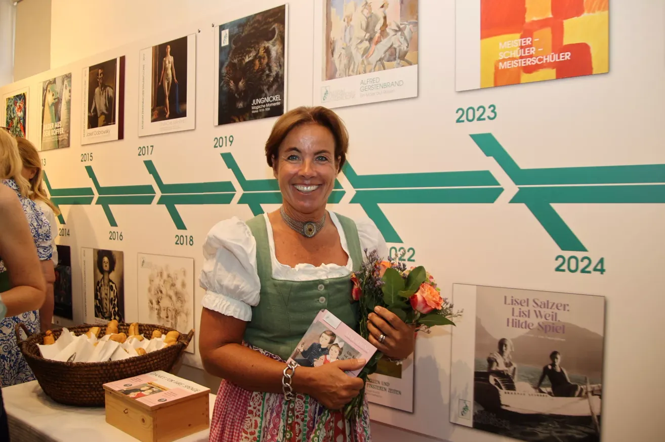 Eine Frau in einem grünen Kleid steht vor einer Wand mit verschiedenen Postern und Kunstwerken. Sie hält einen Blumenstrauß und ein Buch. Die Wand hat eine Zeitlinie der Jahre von 2016 bis 2023.