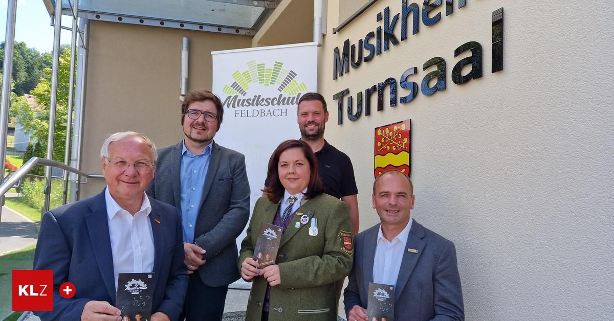 Fünf Personen stehen vor einem Gebäude mit einem Schild, das 'Musikschule Turnsaal' besagt. Sie halten alle Bücher und scheinen für ein Foto zu lächeln. Dahinter befindet sich ein weißes Banner mit dem Text 'Musikschule Feldbach'.