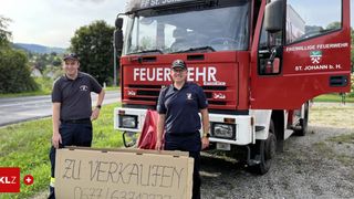 Zwei Feuerwehrleute stehen neben einem roten Feuerwehrauto. Auf der Vorderseite des Autos steht 'Feuerwehr'. Vor ihnen steht ein Karton mit einem Verkaufsschild.