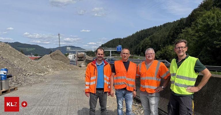 Vier Männer in reflektierenden Westen posieren für ein Foto auf einer Straße mit einem Hintergrund von Bergen und einem blauen Schild.