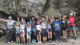 Eine Gruppe von Kindern und Erwachsenen steht vor einer Felsformation. Sie tragen alle Helme und Gurte. Sie heben ihre Hände in die Luft.
