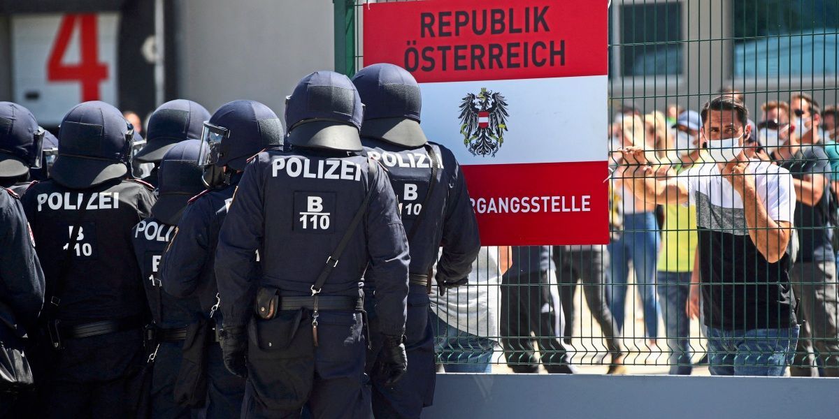 Eine Gruppe von Polizeibeamten steht vor einem Metallzaun. Sie tragen Uniformen und Helme. Ein Schild mit dem Text Republik Osterreich ist am Zaun befestigt. Hinter ihnen stehen Leute.