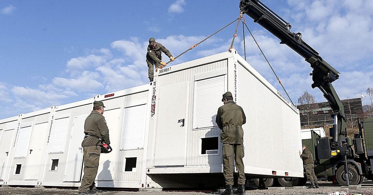 Ein Kran hebt einen weißen mobilen Container, während drei Männer beobachten. Sie tragen Uniformen und Mützen, und einer hält eine Kettensäge. Der Container ist mit Text beschriftet.