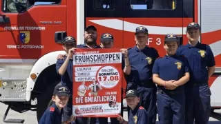 Eine Gruppe Feuerwehrleute in Uniform posiert mit einem Schild für eine lokale Veranstaltung vor einem Feuerwehrauto.