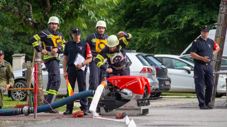 Feuerwehrleute in Uniformen stehen um ein rotes Rosenbauer-Fahrzeug herum, wobei einer der Männer auf die Maschine zeigt. Ein anderer Mann hält ein Papier in der Hand, und sie stehen in der Nähe geparkter Autos.
