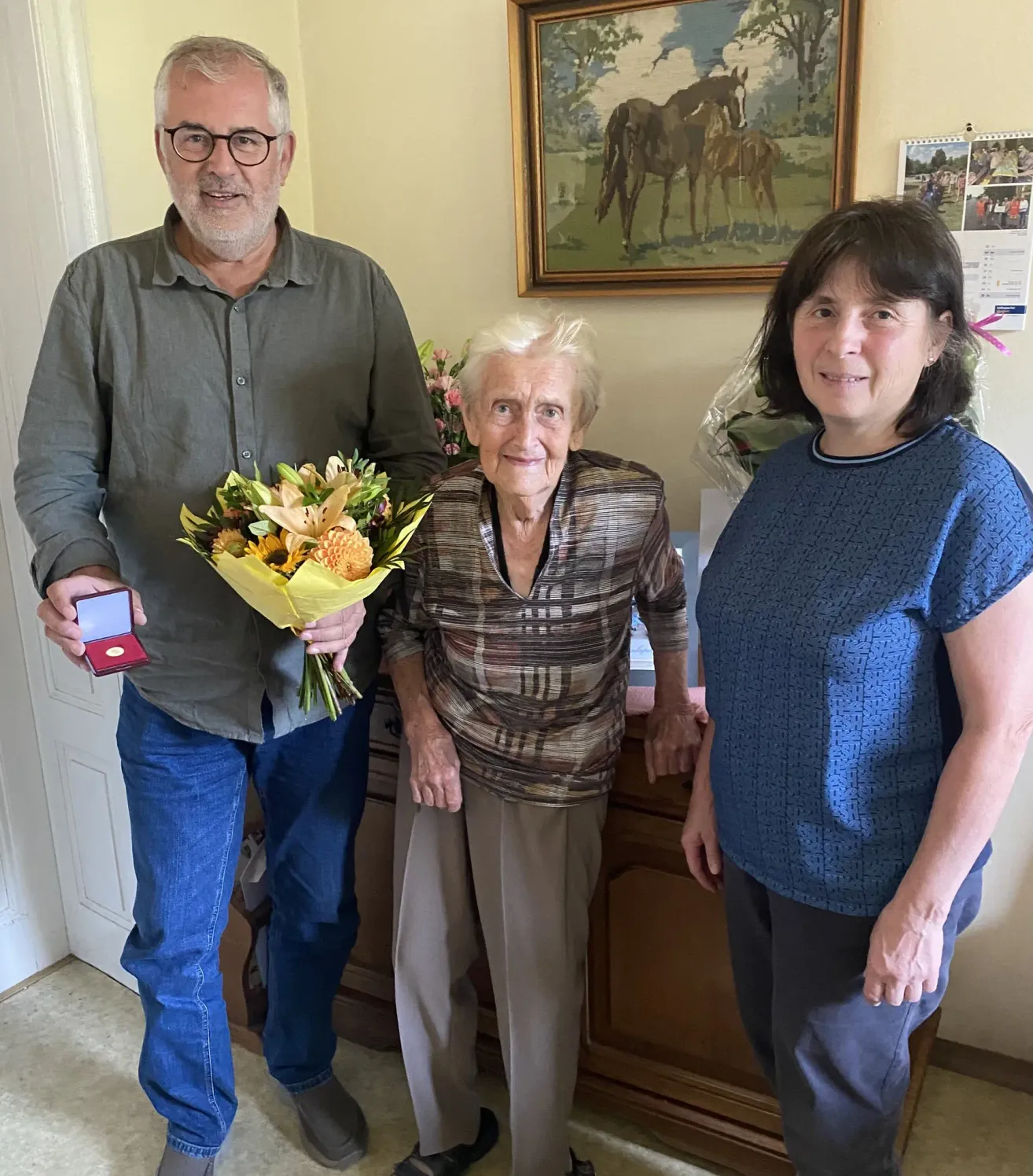Drei Menschen stehen eng beieinander in einem Raum. Ein Mann hält einen Blumenstrauß und eine Medaille. Eine ältere Frau steht zwischen zwei Frauen. Ein Gemälde von Pferden hängt an der Wand.