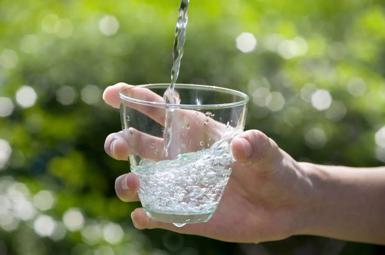 Eine Hand füllt ein klares Glas mit Wasser, das aus einem Wasserhahn gegossen wird. Der Hintergrund ist eine verschwommene grüne Vegetation.