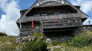 Ein Holzchalet namens Schoenleitenhaus steht auf einem grasbewachsenen Hügel. Es hat einen hölzernen Balkon mit mehreren sitzenden Personen und einem roten Schirm.
