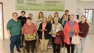 Eine Gruppe älterer Frauen und einige jüngere Frauen posieren für ein Foto in einem Klassenzimmer und halten Tabletts mit Kunst. Ein Banner mit "LFS Warth" und verschiedenen Kursen ist sichtbar.