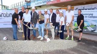 Eine Gruppe von Menschen in formeller Kleidung hält Schaufeln und steht auf einer Schotterfläche, möglicherweise für eine Grundsteinlegungszeremonie. Hinter ihnen befindet sich ein Gebäude mit Bannern und einem Schild.