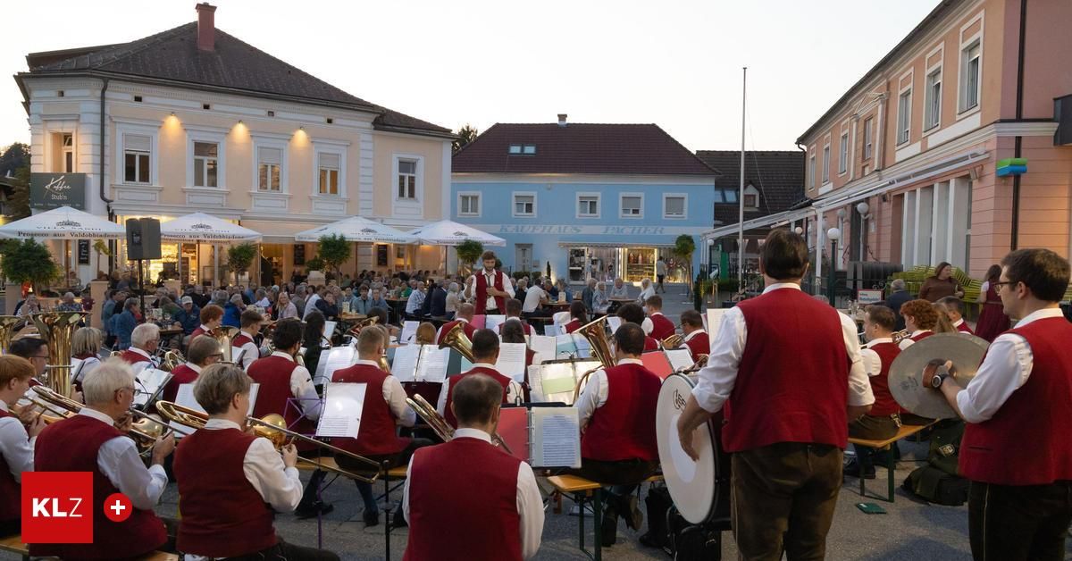 Ein Orchester spielt bei Dämmerung im Freien, mit Musikern in roten Westen, die Instrumente spielen. Zuschauer sitzen vor ihnen, und Gebäude mit Vordächern sind im Hintergrund zu sehen.