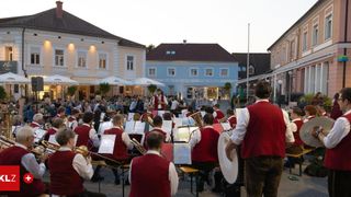 Ein Orchester spielt bei Dämmerung im Freien, mit Musikern in roten Westen, die Instrumente spielen. Zuschauer sitzen vor ihnen, und Gebäude mit Vordächern sind im Hintergrund zu sehen.
