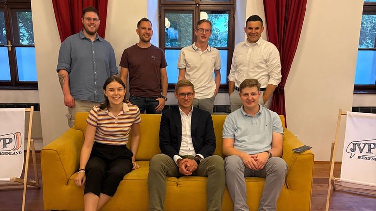 Sieben Personen, sowohl Männer als auch Frauen, versammeln sich in einem Raum, lächeln und posieren für ein Foto. Sie sind lässig gekleidet.
