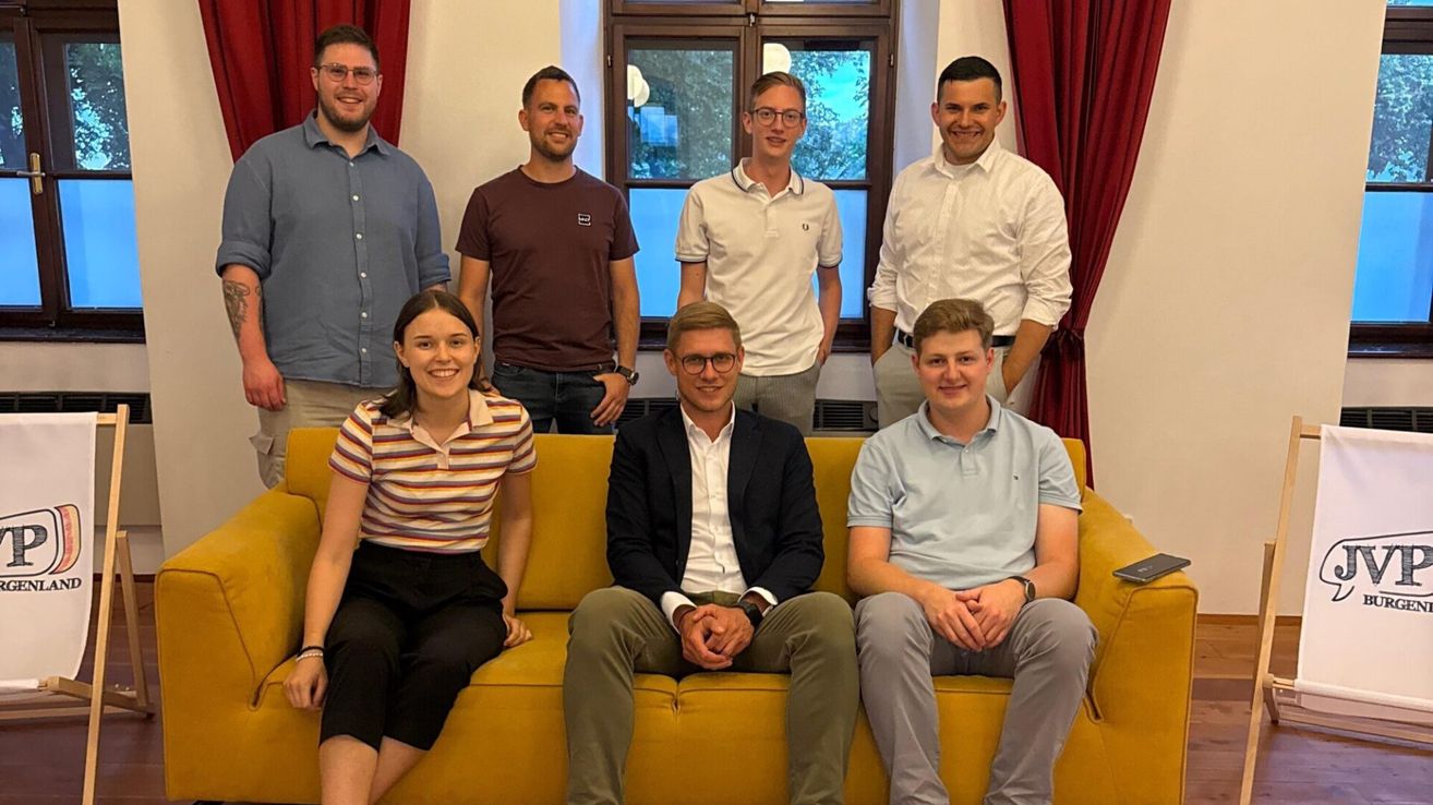 Sieben Personen, sowohl Männer als auch Frauen, versammeln sich in einem Raum, lächeln und posieren für ein Foto. Sie sind lässig gekleidet.