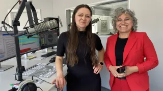Zwei Frauen stehen in einem Studio mit einem Mikrofon und einer Soundtafel. Die Frau auf der linken Seite trägt ein schwarzes Kleid und lächelt. Die Frau auf der rechten Seite trägt einen roten Blazer und hält ein Telefon. Hinter ihnen stehen zwei Computerbildschirme, eine Tastatur und eine Maus.