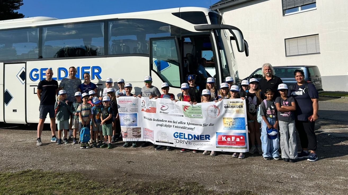 Eine Gruppe von Kindern und Erwachsenen steht vor einem Bus und hält ein Banner. Das Banner hat Text und Logos. Ein Mann steht in der Nähe der Bustür.
