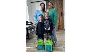 Zwei Frauen und zwei junge Mädchen mit Rucksäcken lächeln und posieren für ein Foto in einem Klassenzimmer.