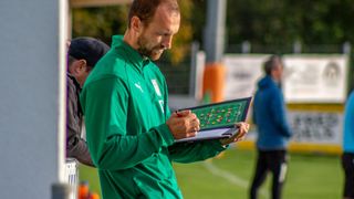 Ein Fußballtrainer in einem grünen Shirt schaut sich ein Tablet mit einer Fußballformation an. Ein anderer Mann steht hinter ihm und ein weiterer läuft in der Ferne.