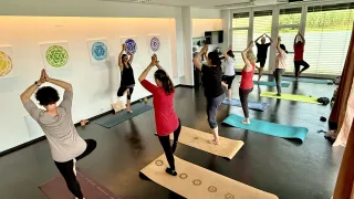 Eine Gruppe von Menschen praktiziert Yoga in Innenräumen. Sie befinden sich in verschiedenen Posen auf Yogamatten. An der Wand hängen drei kreisförmige Kunstwerke.