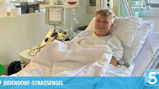 Ein lächelnder Mann liegt in einem Krankenhausbett mit medizinischen Geräten, einschließlich eines IV-Tropfs, neben ihm.
