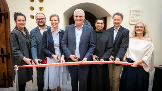 Eine Gruppe von Menschen in formeller Kleidung, die bei einer Schleifchen-Schneidezeremonie stehen. Einige halten Scheren und lächeln. Hinter ihnen ist eine Tafel an der Wand befestigt.