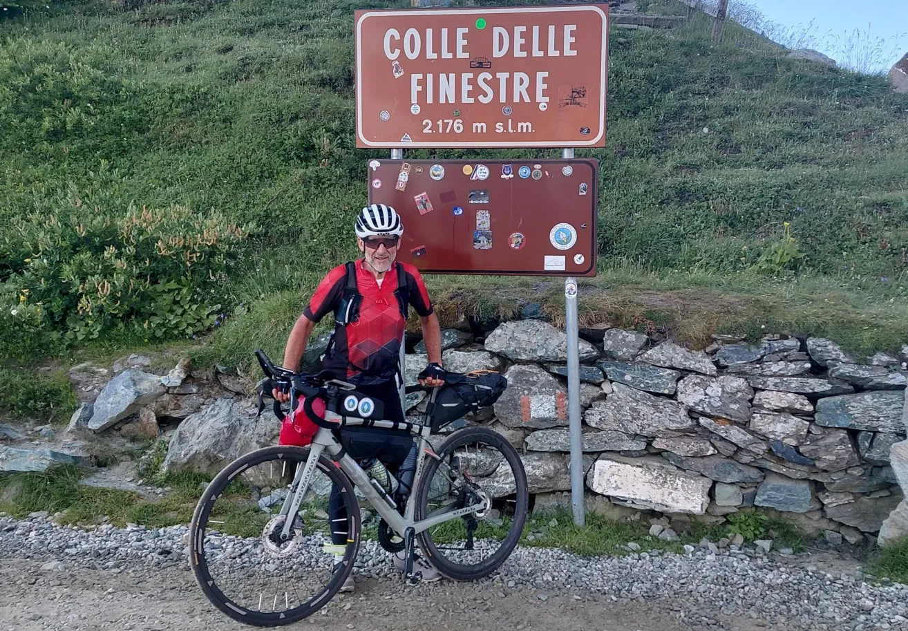 Ein Mann in Fahrradkleidung und mit Helm steht mit seinem Fahrrad vor einem Schild, das 'Colle delle Finestre' besagt. Er befindet sich auf einer Bergstraße mit einer Steinmauer an der Seite.