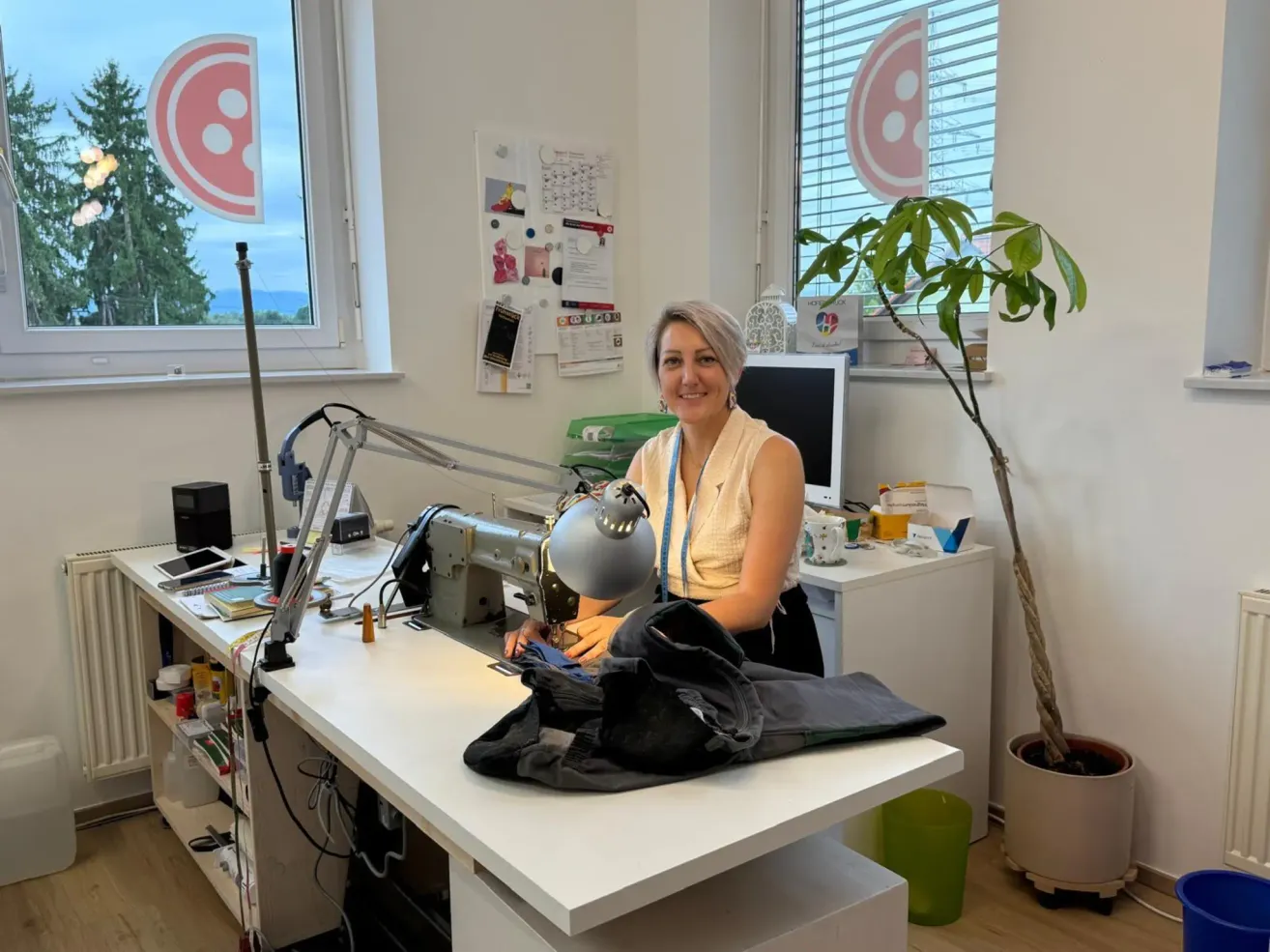 Eine Frau sitzt an einem Schreibtisch in einem hellen Büro, mit einer Nähmaschine und einer Lampe. Eine Pflanze steht in der Ecke und Papiere sind an der Wand angeheftet.