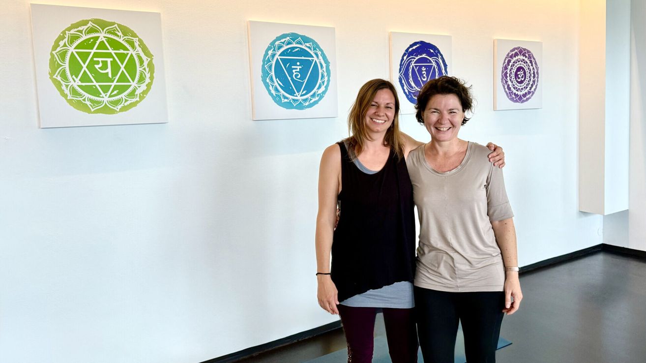 Zwei Frauen stehen lächelnd vor einer Wand mit drei Gemälden. Ein Gemälde hat einen blauen Kreis mit einem Symbol in der Mitte. Die Frauen tragen Freizeitkleidung.