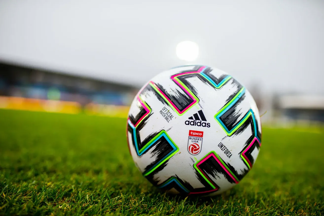 Ein Fußball mit Adidas-Logo und buntem Design liegt auf einem Rasenfeld, mit einem verschwommenen Hintergrund eines Stadions.