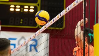 Ein Volleyball mit dem Wort MOOZA in der Mitte eines Volleyballnetzes. Das Netz ist schwarz mit weißer Klebeband. Der Hintergrund ist verschwommen mit einem Anzeigetafel und roter Wand.