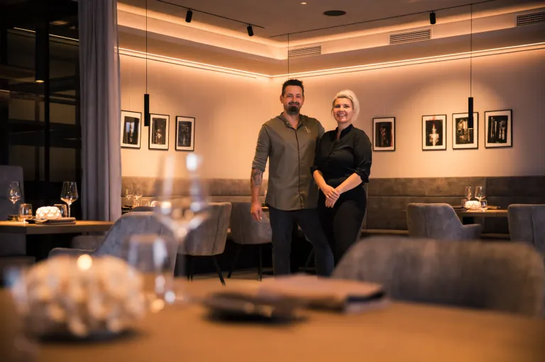Ein Mann und eine Frau stehen in einem modernen Restaurant und lächeln. Sie befinden sich in der Nähe eines Tisches mit Weingläsern und Tellern. Der Raum hat eine minimalistische Einrichtung mit Bilderrahmen an der Wand.