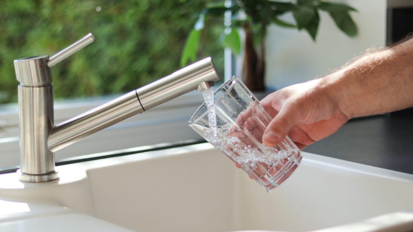 Eine Person füllt ein klares Glas mit Wasser aus einem Edelstahlhahn.