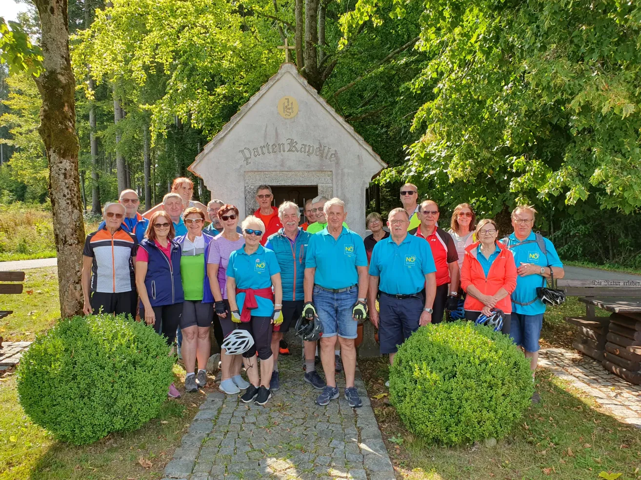 Eine Gruppe von Radfahrern in Radbekleidung posiert für ein Foto vor einer kleinen Kapelle mit dem Wort 'Partenkapelle'. Die Gruppe ist von Grünpflanzen und Bäumen umgeben.