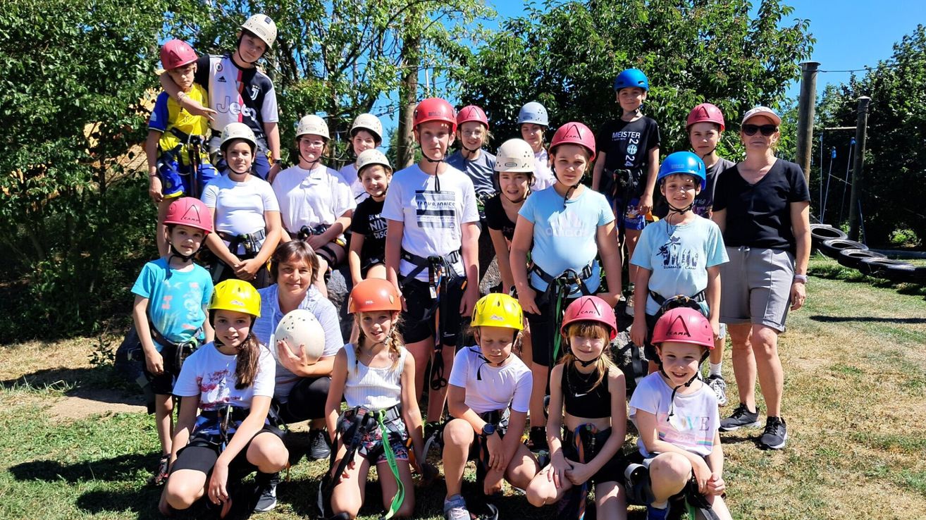Eine Gruppe junger Personen mit Helmen, Gurten und Turnschuhen, die sich für ein Foto versammelt haben, mit Bäumen und blauem Himmel im Hintergrund.