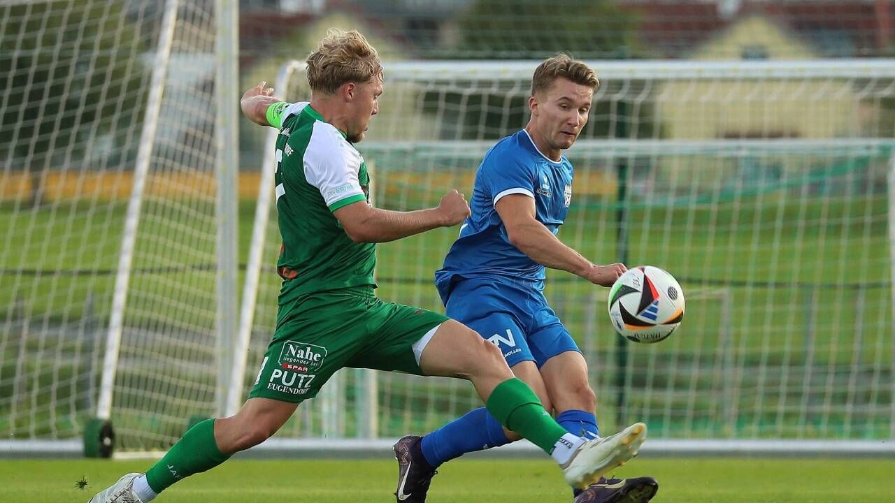 Zwei Fußballer in Aktion, einer in Grün und der andere in Blau. Der Spieler in Grün tritt den Ball, während der Spieler in Blau versucht, ihn zu blocken.