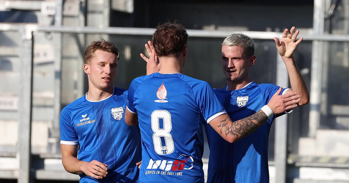 Drei männliche Fußballspieler feiern auf einem Feld. Sie tragen blaue Trikots mit Nummern und Logos. Der Spieler in der Mitte hat eine Tätowierung auf seinem Arm.
