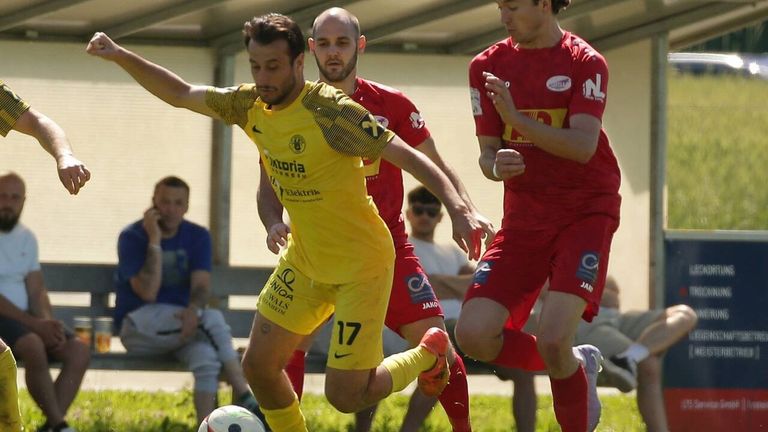 Drei Fußballspieler sind auf dem Feld in Aktion. Der Spieler im gelben Trikot jagt dem Ball hinterher, während die beiden Spieler in roten Trikots versuchen, ihn zu blockieren. Im Hintergrund sitzen einige Leute auf der Bank.