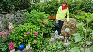 Eine ältere Frau mit Brille und Schal steht in einem Garten. Hinter ihr ein großer Fels und ein Holzschild mit der Aufschrift 'Traumgarten'. Der Garten ist voller Blumen und Pflanzen.